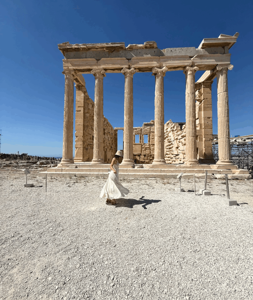 Person in a white dress walking before ancient Greek columns under a clear blue sky—timeless architecture as a metaphor for strong creative foundations.
